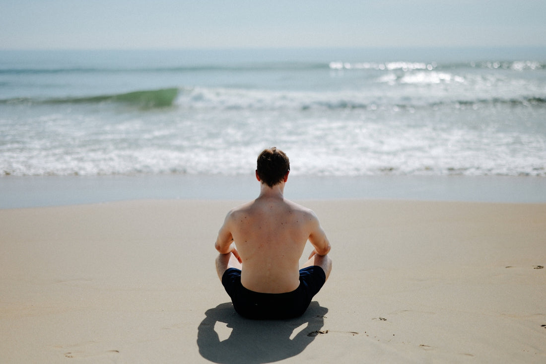 Man meditating on a sandy beach facing the ocean.